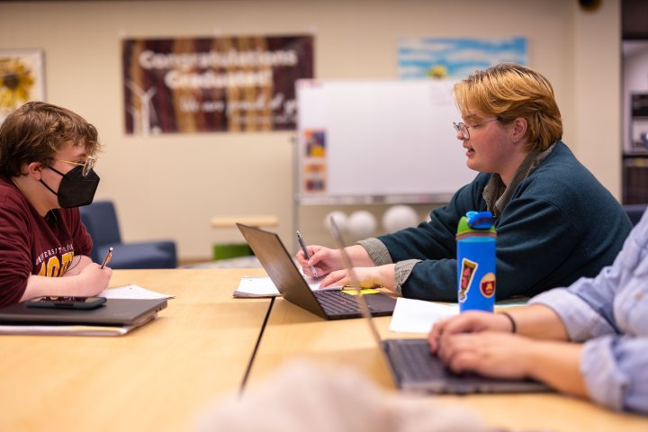 Two students sit at a table in a library setting, engaged in discussion and working on laptops. One student wears a mask, taking notes, while the other gestures as they speak. A whiteboard and banners can be seen in the background.